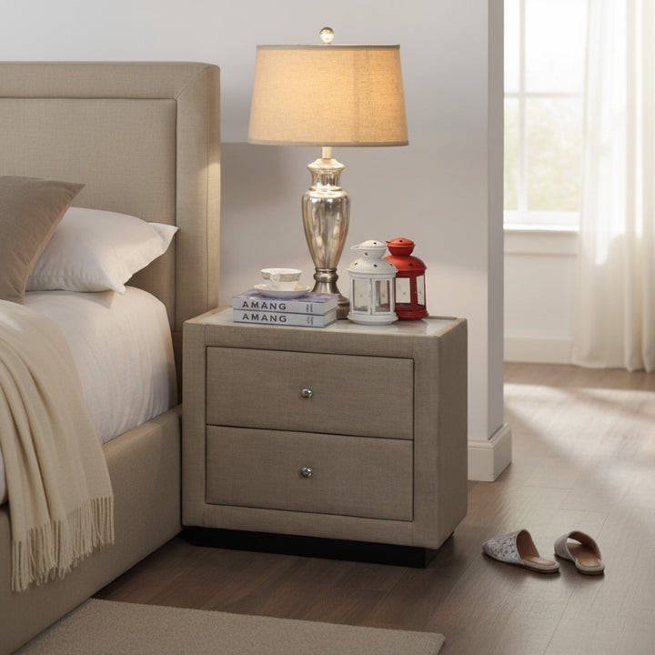 Modern beige upholstered nightstand with drawers, lamp, books, and lanterns beside bed