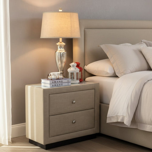 Modern beige nightstand with two drawers, table lamp, books, and decor beside upholstered bed