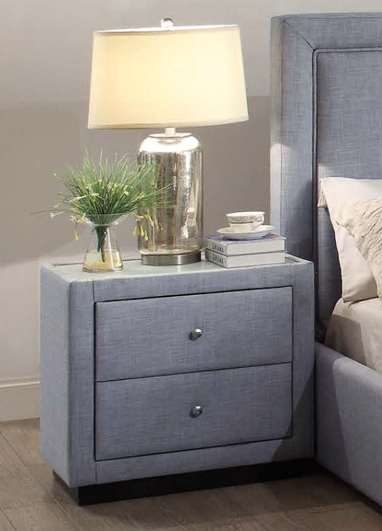 Modern grey upholstered nightstand with two drawers, lamp, books, and plant decor