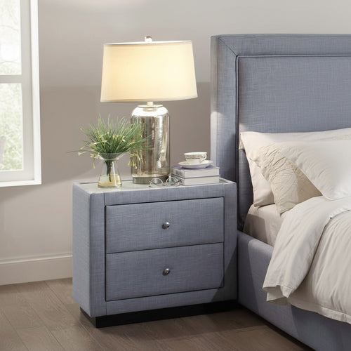 Modern grey upholstered nightstand with two drawers beside a matching bed, topped with a table lamp, books, and decor in a bright bedroom.