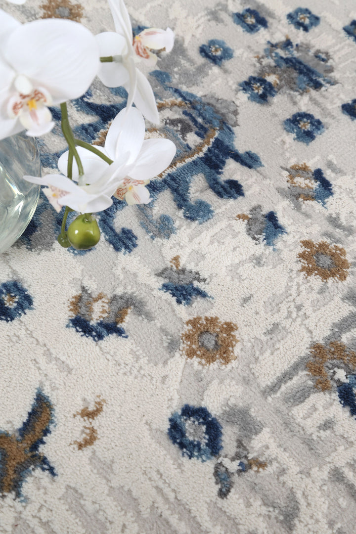 Close-up of patterned area rug with blue, beige, and gray floral design and white orchid flowers in a glass vase.