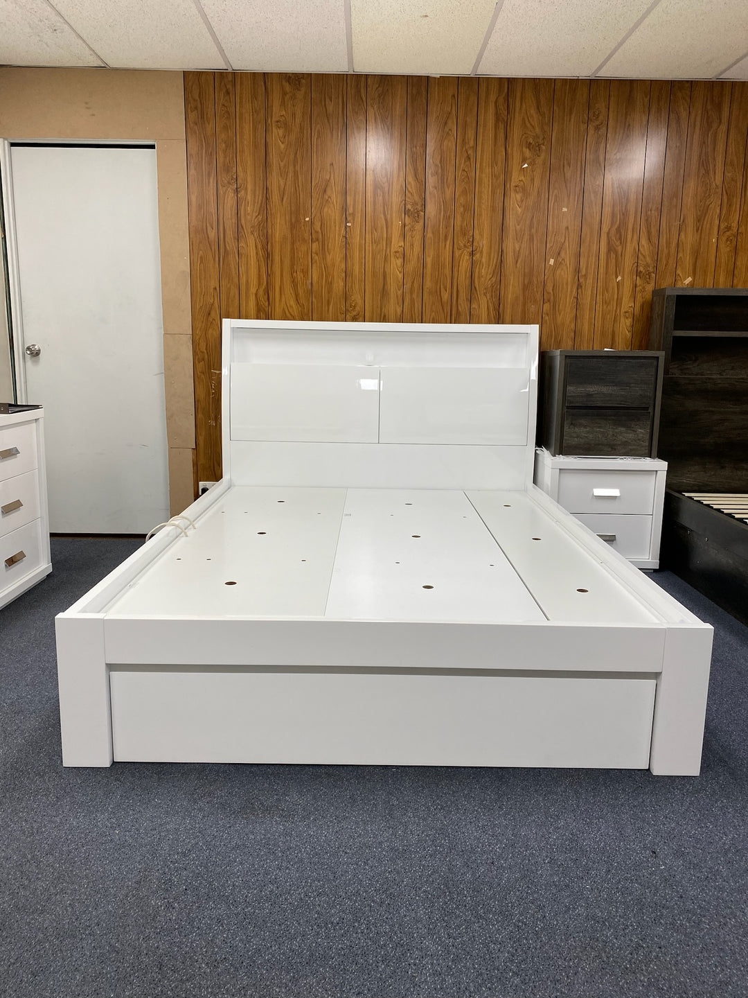 Modern white platform bed with storage, displayed in a furniture showroom with wood panel walls.
