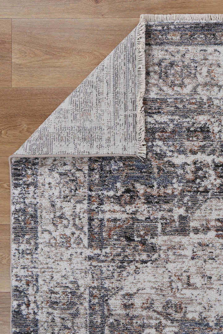 Distressed grey and blue area rug with fringe on wooden floor, corner flipped to show backing