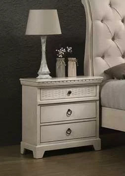 White wooden bedside table with three drawers, lamp, and decor in a bedroom setting