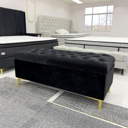 Black tufted storage ottoman with gold legs in modern furniture showroom with upholstered beds
