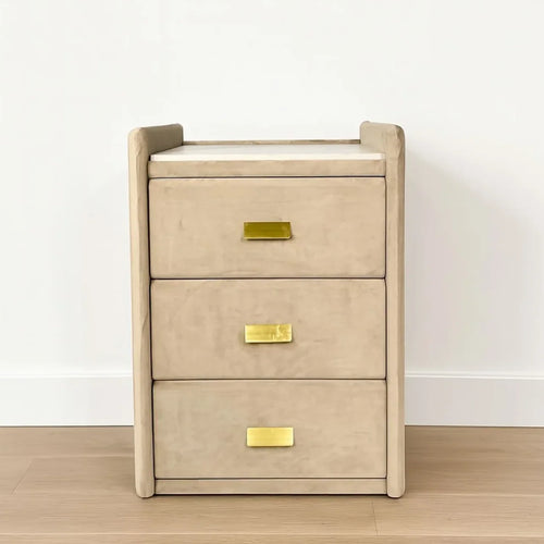 Beige bedside table with three drawers and gold handles on a wooden floor.