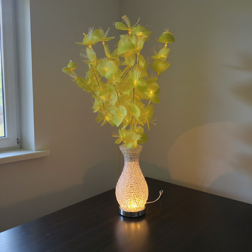Glowing yellow flower lamp with textured vase base on dark table near window